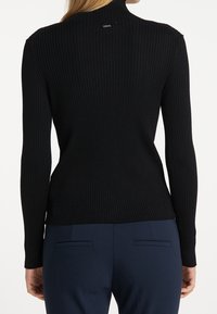 Zwarte geribbelde turtleneck sweater met lange mouwen, uitgerust met een getailleerd ontwerp en een klein logo-label op de achterkant van de kraag. Gecombineerd met marineblauwe broeken.