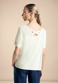 Bílá halenka s krátkým rukávem se vzadu s výřezem a kříženými ramínky, kombinovaná s tmavě modrými širokými džínami. Hladký materiál.