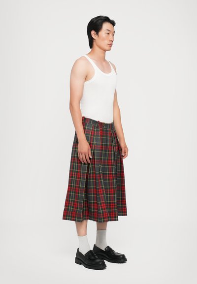 Rot- und grünkariertes Kilt, plissiertes Design, kombiniert mit einem weißen Tanktop und schwarzen Schuhen. Sichtbarer Gürtel und Socken. Minimalistischer Hintergrund.