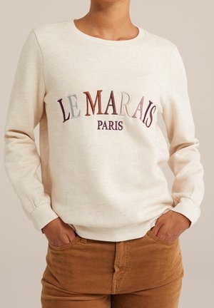 Beige crewneck sweatshirt with multicolored "LE MARAIS PARIS" lettering, worn with brown corduroy pants, hands in pockets.