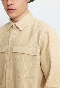 Beige corduroy overhemd met een kraag, twee borstzakken en knoopsluiting. Heeft een verticale ribstructuur. De knopen zijn lichtbeige.