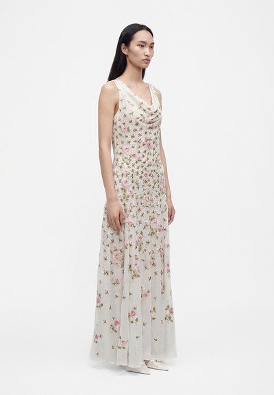 Femme aux longs cheveux foncés portant une robe blanche longue sans manches avec une broderie florale rose et des chaussures blanches pointues, debout de profil.