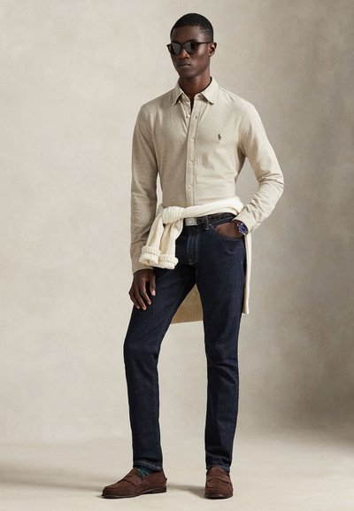 Camisa bege de manga comprida com fecho de botões, jeans escuros, mocassins castanhos e óculos de sol. Suéter branco amarrado à cintura. Design simples.