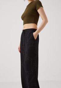 Olivově zelený crop top s krátkými rukávy, který je kombinován s černými kalhotami s vysokým pasem, jež mají kontrastní béžové geometrické prošívání a boční kapsy.