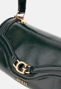 Bolso de mano de charol verde con un acento de logo dorado y diseño curvado. Presenta una textura suave y costuras decorativas a lo largo del borde.