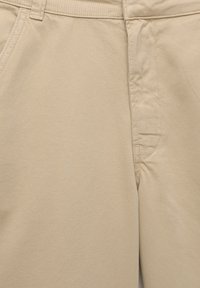 Beige színű pamut nadrág, egyenes fazonnal, látható varrással és oldalsó zsebekkel. Az anyag sima, kissé strukturált felületű.