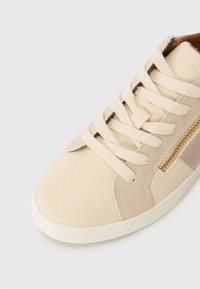 Beige skinnesneaker med en glatt tekstur, som har kryss-søm detaljer, flate skolisser og en dekorativ glidelås på siden.
