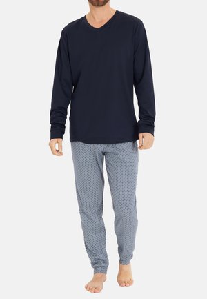 Mann trägt ein marineblaues Langarm-V-Neck-Shirt und hellblaue gemusterte Pyjamahosen, steht barfuß auf einfarbigem Hintergrund.