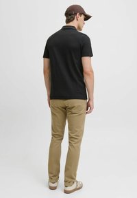 Zwarte polo, kaki slim-fit broek en beige sneakers. Het model staat gedeeltelijk met de rug naar de camera, met een bruine pet op. Eenvoudige, casual outfit.