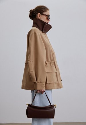 Frau trägt eine beige Jacke mit hohem Kragen, helle Hose, Sonnenbrille und hält eine dunkelbraune Handtasche vor einem einfarbigen Hintergrund.