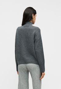 Γκρι πλεκτό πουλόβερ με υψηλό γιακά, ελαφρώς φαρδιά εφαρμογή και ριμπ τελείωμα. Φορεμένο με ανοιχτό γκρι, μαλακά υφασμένα παντελόνια σε casual περιβάλλον.