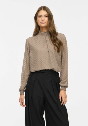 Femme portant un chemisier taupe texturé à col haut et manches longues, associé à un pantalon noir large taille haute sur fond blanc.