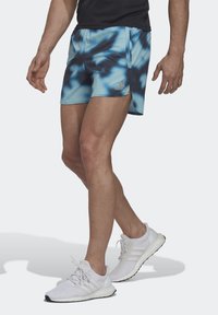 Shorts bleu et noir en tie-dye avec une taille élastique, ornés d'un logo. Associés à des chaussures de sport blanches et un haut noir.
