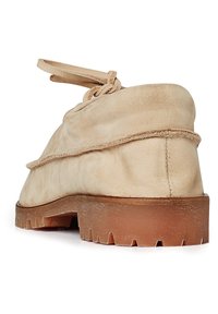 Zapato de ante beige con una textura suave, suela de goma plana y un detalle de cordones en bucle en la parte trasera. Presenta un tacón ligeramente elevado.