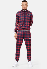 Tracksuit made of smooth, red and navy plaid fabric. Features a zip-up jacket with striped sleeves and matching pants. White branding logo displayed.