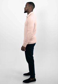 Helles rosa Langarm-Polohemd mit Kragen, kombiniert mit dunkelblauen Slim-Fit-Hosen und schwarzen Slippern. Glatte Textur.