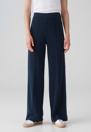 Wide-leg navy pants with a high elastic waistband, side pleats, and a smooth texture. Paired with white athletic shoes.