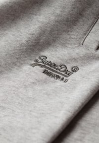 La tela gris presenta el logo "SuperDry" bordado en un gris más oscuro. El material es suave con una textura sutil. La vista en primer plano resalta los detalles de la costura.
