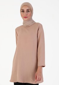 Langærmet beige tunika lavet af blødt stof, der har en høj halsudskæring og lige snit. Modellen bærer en matchende hijab.
