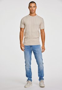 Μπεζ κοντό μανίκι πουλόβερ με κάθετη ριμπ υφή, σε συνδυασμό με ανοιχτό μπλε τζιν και λευκά αθλητικά παπούτσια, σε έναν απλό φόντο.