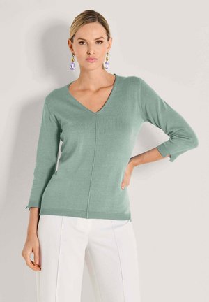 Vrouw draagt een saliegroene V-hals trui met driekwart mouwen en witte broek, poserend met één hand op de heup tegen een neutrale achtergrond.
