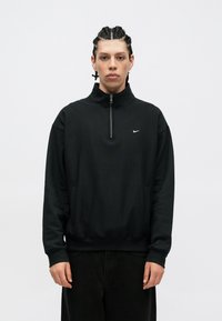 Fekete Nike pulóver, puha anyagból készült, magas nyakkal, félig cipzárral és elülső zsebekkel rendelkezik. Egyszerű design, minimális márkázással.