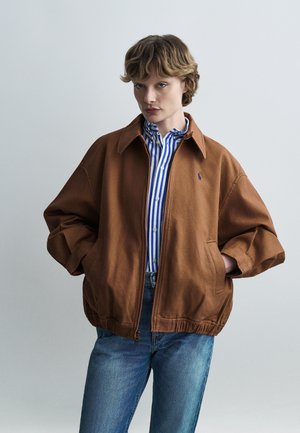 Άτομο που φοράει καφέ ζακέτα με φερμουάρ πάνω από μπλε-λευκό ριγέ πουκάμισο με κουμπιά και μπλε τζιν, στέκεται με τα χέρια στις τσέπες.