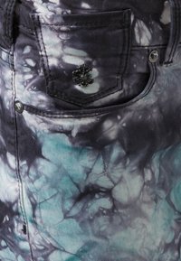 Jean en denim avec un design tie-dye dans des tons de gris et de teal, agrémenté d'un bouton décoratif en forme de fleur noir et de détails sur la poche avant.