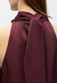 Djupt vinröd halterneck-top med draperad halsbindning. Tyget är mjukt och något genomskinligt, med en avslappnad passform och subtil textur.