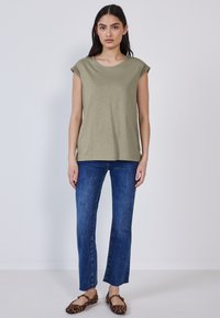 Top senza maniche verde oliva con scollatura rotonda e pizzo lungo le spalle, abbinato a jeans blu e ballerine con stampa animale. Sfondo semplice.