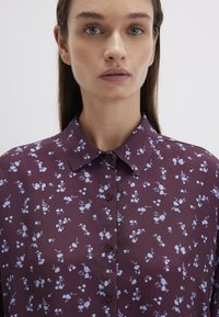 Burgundy blouse with a floral pattern in light blue, featuring a button-down front and collar. Made from a smooth, lightweight fabric.