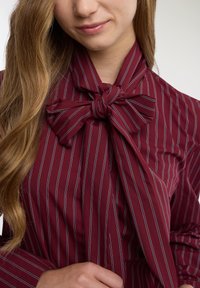 Blusa color bordeaux con righe verticali bianche, caratterizzata da un dettaglio a fiocco sul colletto e da una texture liscia del tessuto. Silhouette aderente.