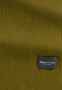 Oliwkowa zielona dzianina prążkowana z czarną prostokątną metką Marc O’Polo przyszytą na tkaninie, pokazująca nazwę marki i napis "EST. IN STOCKHOLM".