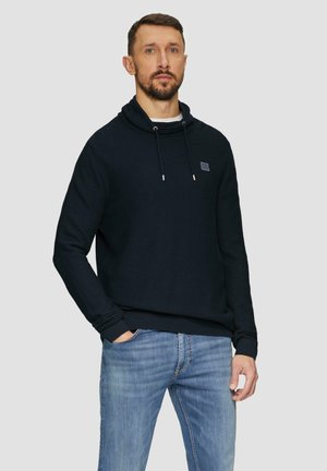 Marineblauwe fleece hoodie met een hoge kraag, verstelbaar koord, voorzak en klein logo-patch. Gecombineerd met lichtblauwe jeans.