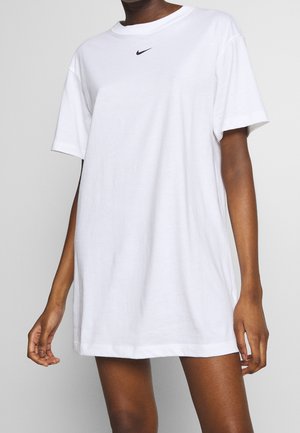 Persoon draagt een loszittende witte Nike T-shirtjurk met korte mouwen, staand tegen een effen lichtgrijze achtergrond.