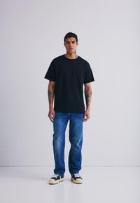 Črna bombažna majica s kratkimi rokavi, v kombinaciji z dodelanimi modrimi jeans hlačami in črnimi športnimi čevlji z belimi detajli, stojijo na beli podlagi.