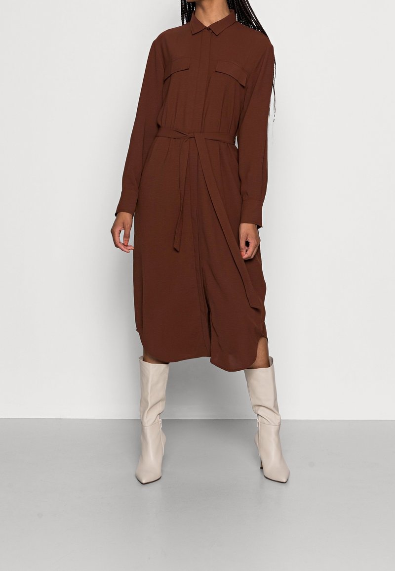 Robe brune ceinturée avec un col, des manches longues et deux poches avant, associée à des bottes à talons beiges jusqu'aux genoux.