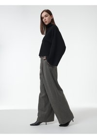 Vrouw in een losse zwarte coltrui, brede grijze broek en zwarte pumps met spitse neuzen, staande tegen een witte achtergrond.