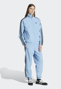 Light blue athletic tracksuit with black side stripes, zippered jacket, elastic cuffs, and matching pants. Black sneakers complete the look.