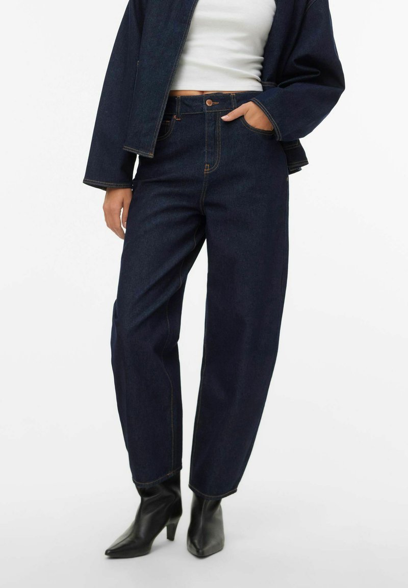 Tamsiai mėlynos aukšto liemens džinsinės kelnės tiesiomis kojomis, kontrastinėmis siūlėmis ir penkiomis kišenėmis, derinamos su juodais kulniukais su smailiais pirštais.