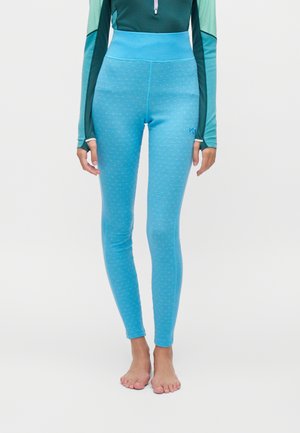 Persona con leggings de talle alto de color azul brillante con un patrón sutil, de pie descalza contra un fondo claro y sencillo.
