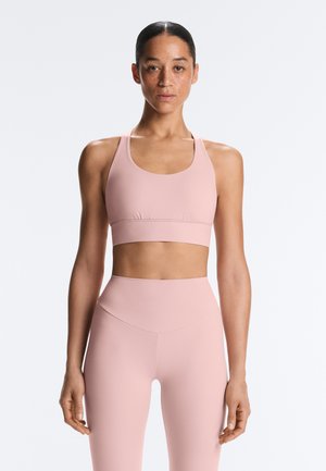 Nő visszafogott hajjal, halvány rózsaszín sportmelltartóban és magas derekú leggingsben, fehér háttér előtt áll, szemben a nézővel.