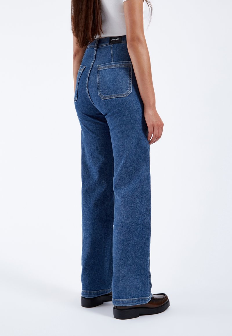 Femme portant un jean bleu taille haute à jambe droite, un haut blanc et des chaussures à plateforme noires, debout, dos à la caméra sur fond blanc.