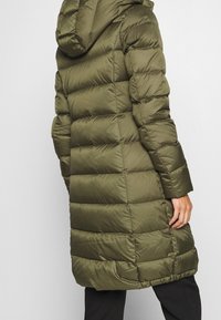 Cappotto puffer verde oliva con cappuccio, caratterizzato da un design trapuntato, di lunghezza intermedia e dalla texture liscia. Include una vita leggermente sfinata e maniche lunghe.