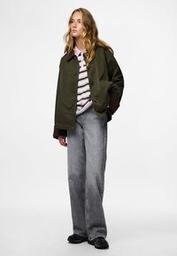 Olive rohelise värvi suurendatud jakk pruunide aksentiidega, kihiliselt seljas roosa ja valge triibuline särk, koos laia jala hallide teksade ja mustade tossudega.