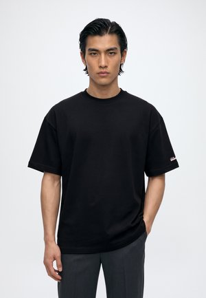 Un homme porte un T-shirt noir uni et un pantalon foncé sur un fond clair de studio.