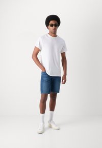 T-shirt blanc, short en denim bleu et baskets blanches avec des accents beige. Le modèle porte des lunettes de soleil vertes et se tient avec une main dans sa poche.