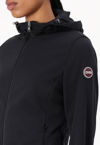 Svart softshell-jacka med dragkedja framtill, samlad krage och Colmar-logotyp på vänster arm. Har en slät, strukturerad tyg.