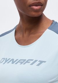 Κοντινό πλάνο ενός ατόμου που φοράει ανοιχτό μπλε αθλητική φανέλα με γκρι πάνελ στους ώμους και τη λέξη «DYNAFIT» τυπωμένη στο στήθος.