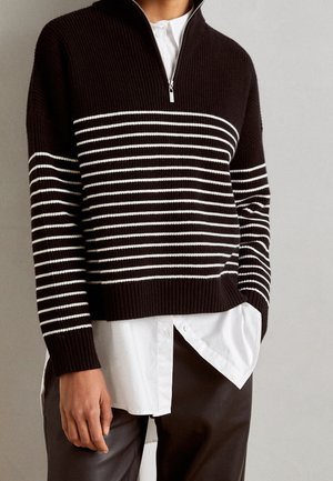 Person iført en sort ribbet kvart-lynlås sweater med hvide vandrette striber over en hvid skjorte med knapper og mørke bukser.